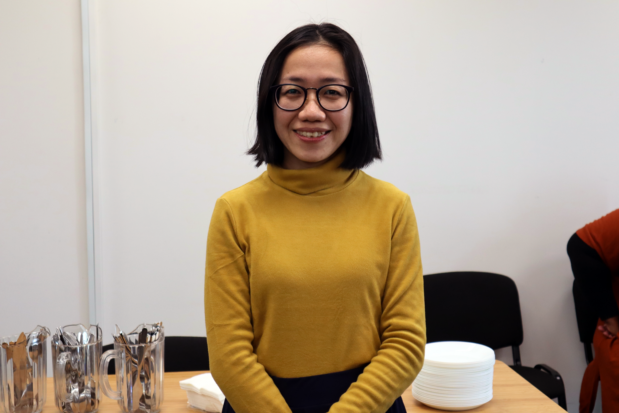 mid-shot of a young woman wearing a yellow jumper and glasses smiling at the camera.